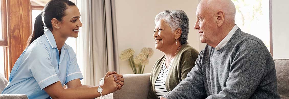 Female caregiver chats with a senior couple at home sitting on sofa.